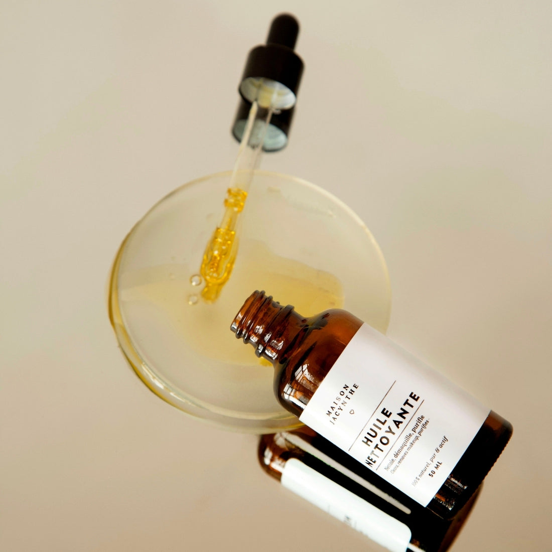 Bouteille et pipette en vitre couchées avec une flaque de l'Huile nettoyante de Maison Jacynthe